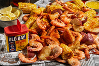 Old Bay Seasoning next to a seafood boil with shrimp, corn on the cob, red potatoes, and lemon wedges